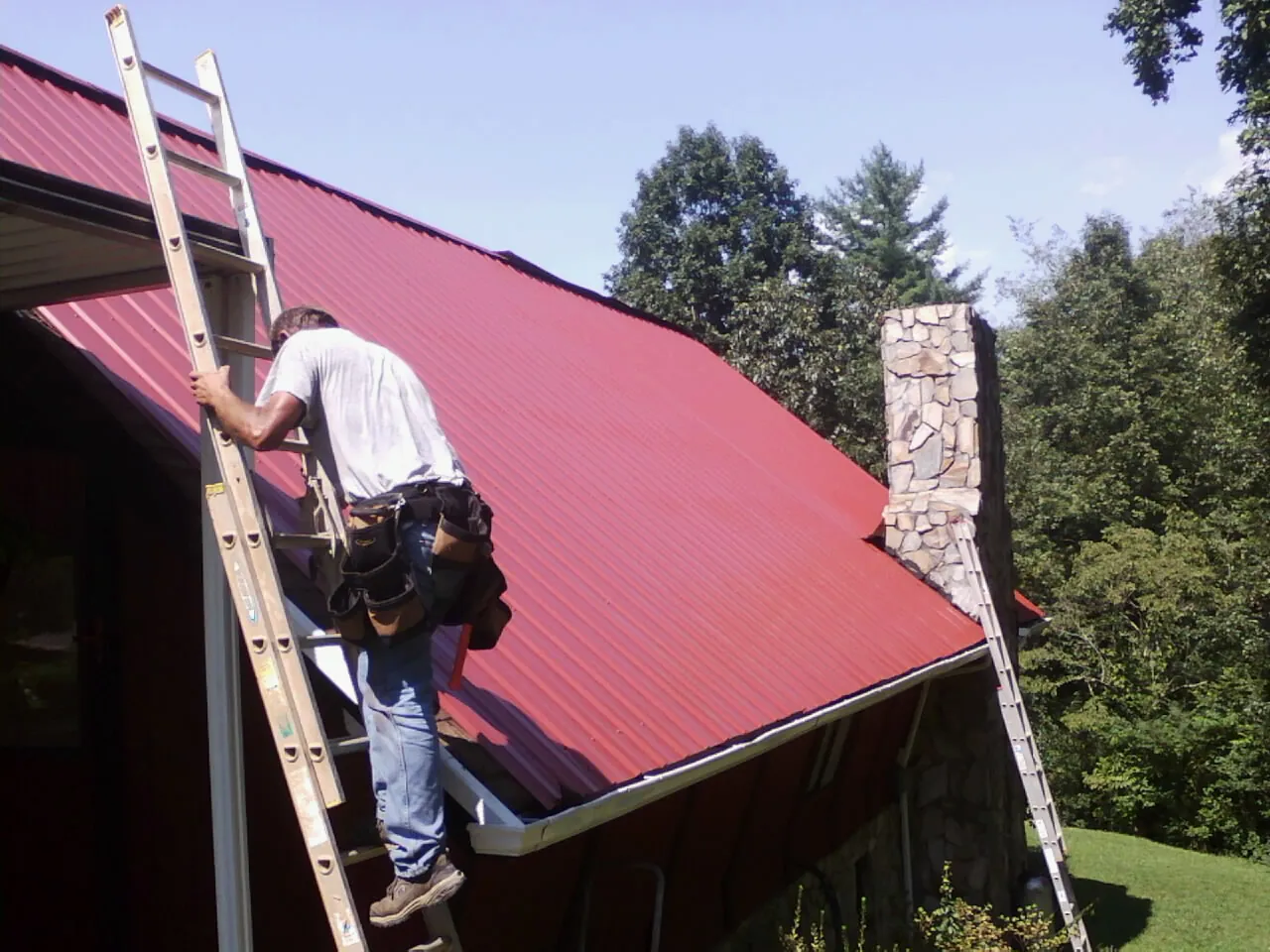 Roofer on a ladder beside a red metal roof and stone chimney — Soffit & Fascia Repair by Roofing Brothers in Mendota